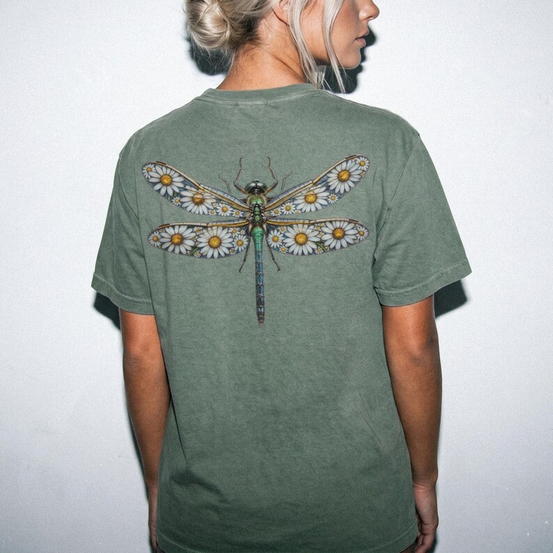 May include: Olive green t-shirt featuring a detailed dragonfly design on the back. The dragonfly's wings are adorned with white daisies and yellow centers. The insect's body is a mix of green, blue, and brown hues.