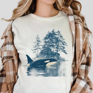 May include: Cream-colored t-shirt featuring a detailed illustration of an orca swimming in the ocean near a small island with trees. The orca is black and white, and the trees are a muted blue. The shirt is worn with a brown and tan plaid jacket.
