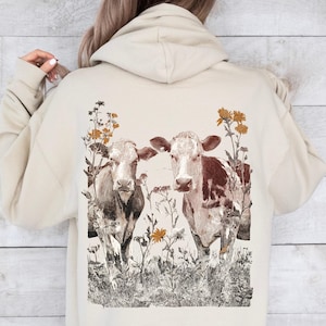 May include: A beige hoodie featuring a sepia-toned illustration of two cows standing in a field of wildflowers. The cows have brown and white markings. The hoodie is made of a soft material.