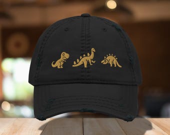 May include: Black distressed baseball cap featuring three gold embroidered dinosaur designs. The dinosaurs include a T-Rex, a Brontosaurus, and a Stegosaurus. The cap has a curved brim and an adjustable strap.