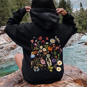 May include: Black hoodie featuring a colorful floral design on the back. The design includes various flowers in shades of red, pink, orange, white, and blue. The hoodie has a drawstring hood and long sleeves.