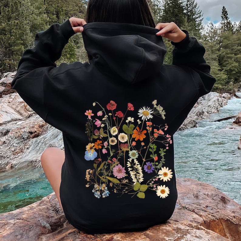 May include: Black hoodie featuring a colorful floral design on the back. The design includes various flowers in shades of red, pink, orange, white, and blue. The hoodie has a drawstring hood and long sleeves.