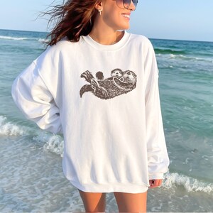 May include: White sweatshirt with a brown graphic of two sea otters cuddling. The otters are facing each other and the one on top has its arms wrapped around the other otter.