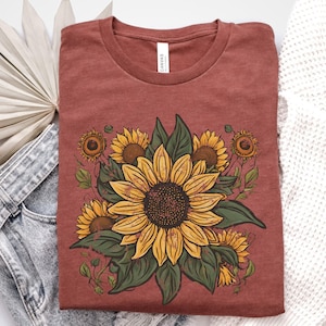 May include: A rust-colored t-shirt featuring a detailed sunflower design. The graphic includes several sunflowers in various stages of bloom, with yellow petals, brown centers, and green leaves. The shirt is made of soft material.