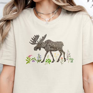 May include: Beige t-shirt featuring a detailed illustration of a moose with large antlers, walking among colorful wildflowers. The design is in shades of brown and green, creating a nature-inspired aesthetic. The shirt is a neutral color.