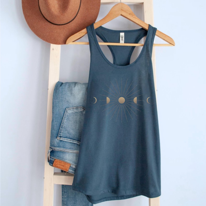 May include: A teal tank top with a gold sun and moon phase design. The tank top is hanging on a wooden hanger, with a pair of blue jeans and a brown hat in the background. The tank top is made of a soft material.