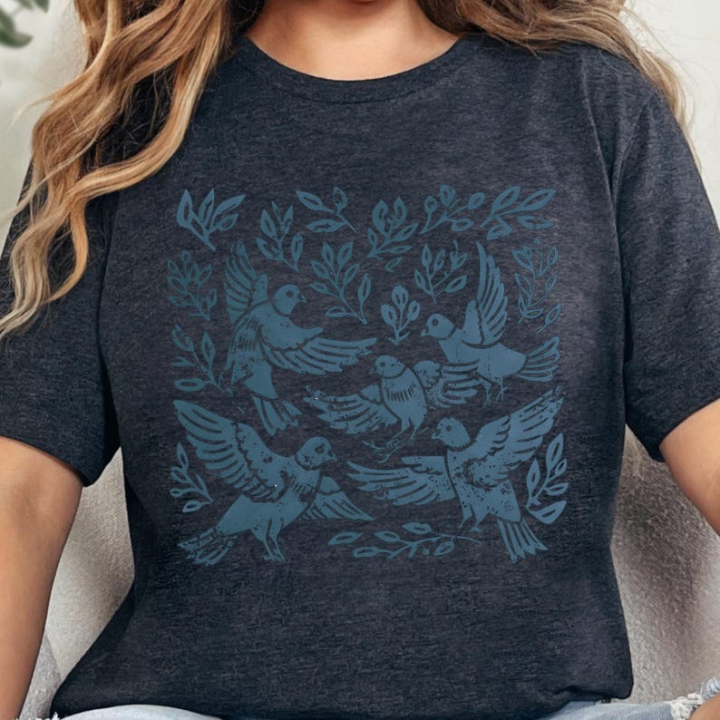 Pu&ograve; includere: T-shirt grigio scuro con un'illustrazione color teal di uccelli e fogliame. Il design include diversi uccelli con le ali spiegate, circondati da foglie e rami. La maglietta ha maniche corte e girocollo.