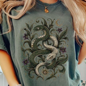 Op de afbeelding: Een saliegroen t-shirt met een gedetailleerde grafische print van twee in elkaar verstrengelde slangen, omringd door donkergroen gebladerte, kleine paarse bloemen, een maansikkel en een zonnesymbool, gecentreerd op de borst van het kledingstuk.