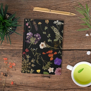 May include: A black journal with a pressed flower design. The flowers are in various colors, including purple, pink, white, and yellow. The journal is closed and lying flat on a wooden surface.