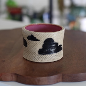 May include: A cylindrical ceramic cup with a textured beige exterior featuring black painted cloud shapes and diagonal etched lines. The interior of the cup is finished in a solid, smooth pink glaze, creating a contrast with the outer surface.