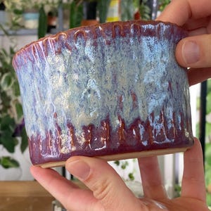 May include: A handmade ceramic planter with a unique glaze. The planter features a cylindrical shape with a textured surface. The colors include shades of blue, purple, and cream, creating a marbled effect. The rim has a ridged design.