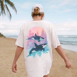 May include: White t-shirt featuring a graphic of two orcas in a colorful sunset scene. The orcas are black and white, with a backdrop of pink, purple, and blue hues. The shirt is a casual, oversized style.