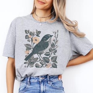 May include: A light gray t-shirt featuring a detailed illustration of a bird perched among leaves and flowers. The bird is primarily dark gray with a white throat, and the flowers are a soft peach color. The design is centered on the shirt.