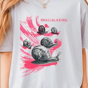 May include: A light gray t-shirt featuring a graphic design of several snails in black and white, with pink brushstroke accents. The word "SNAILBLAZING" is printed in pink above the snails.