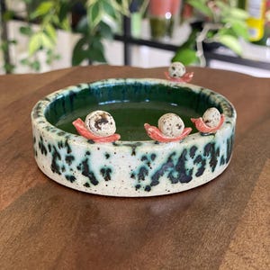 May include: A ceramic bowl with a speckled white and teal exterior and a deep green interior. The bowl is decorated with three small, speckled eggs resting on red, leaf-shaped accents. The bowl is round and sits on a wooden surface.
