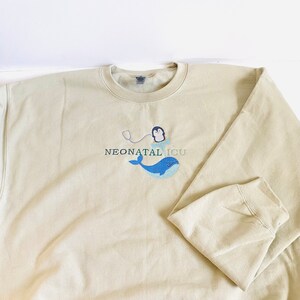 Pode incluir: Camisola bege com o texto bordado "NEONATAL ICU". O design apresenta uma baleia azul, um pinguim com um estetosc&oacute;pio e o texto em cor azul-petr&oacute;leo. A camisola tem gola redonda e mangas compridas.