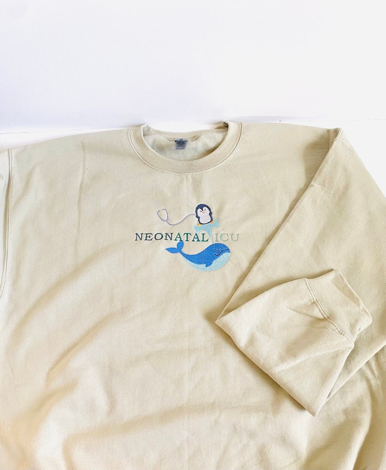 Pode incluir: Camisola bege com o texto bordado "NEONATAL ICU". O design apresenta uma baleia azul, um pinguim com um estetosc&oacute;pio e o texto em cor azul-petr&oacute;leo. A camisola tem gola redonda e mangas compridas.