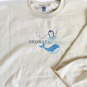 Pode incluir: Camisola bege com o texto bordado "NEONATAL ICU". O design apresenta um pinguim com estetosc&oacute;pio, uma baleia azul e uma nuvem. A camisola tem mangas compridas e gola redonda.