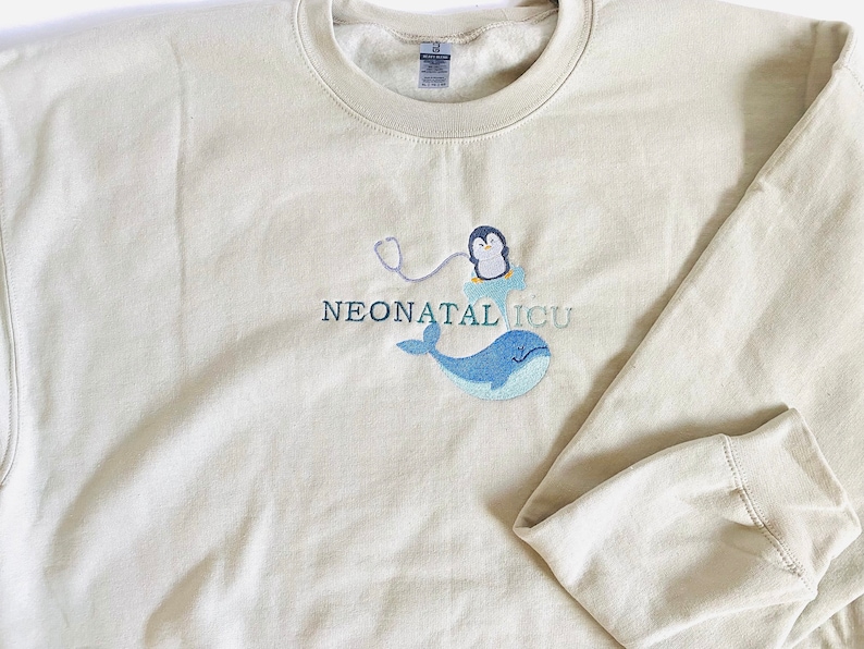Pode incluir: Camisola bege com o texto bordado "NEONATAL ICU". O design apresenta um pinguim com estetosc&oacute;pio, uma baleia azul e uma nuvem. A camisola tem mangas compridas e gola redonda.