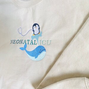 Pode incluir: Camisola bege de gola redonda com design bordado. Apresenta um pinguim com um estetosc&oacute;pio, uma baleia azul e as palavras "NEONATAL ICU" em verde-azulado. A camisola &eacute; feita de um material macio.