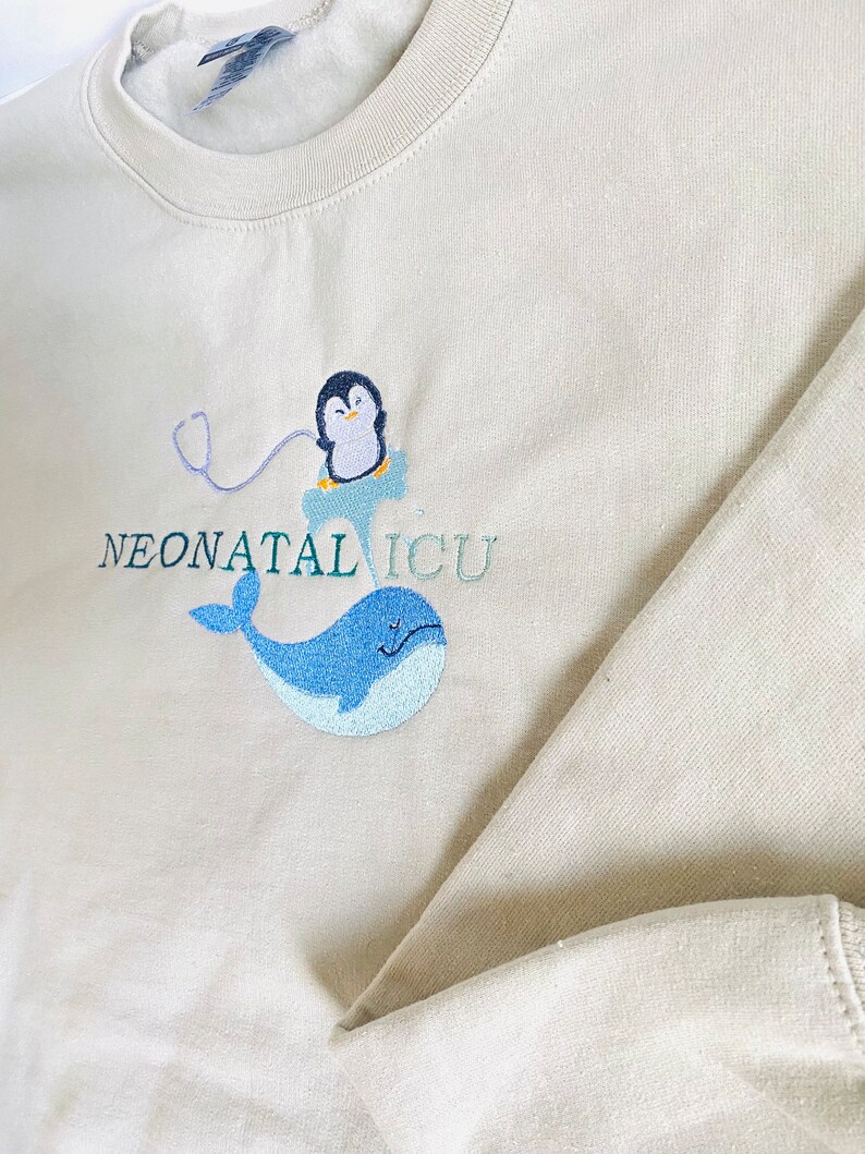 Pode incluir: Camisola bege de gola redonda com design bordado. Apresenta um pinguim com um estetosc&oacute;pio, uma baleia azul e as palavras "NEONATAL ICU" em verde-azulado. A camisola &eacute; feita de um material macio.