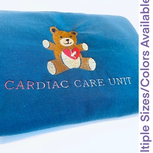 Puede incluir: Una sudadera azul marino con un oso de peluche bordado sosteniendo un corazón rojo. El texto "CARDIAC CARE UNIT" está bordado en hilo rojo. La imagen también incluye el texto "Multiple Sizes/Colors Available" en una columna vertical.