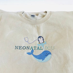 Pode incluir: Camisola bege de gola redonda com bordado. O design inclui uma baleia azul, um pinguim com estetosc&oacute;pio e o texto "NEONATAL ICU" em verde e roxo. O bordado est&aacute; na frente.