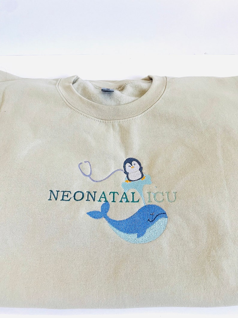 Pode incluir: Camisola bege de gola redonda com bordado. O design inclui uma baleia azul, um pinguim com estetosc&oacute;pio e o texto "NEONATAL ICU" em verde e roxo. O bordado est&aacute; na frente.
