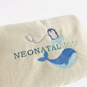 Pode incluir: Um cobertor bege claro bordado com as palavras "NEONATAL ICU". O design inclui uma baleia azul, um pinguim com um jaleco m&eacute;dico e um estetosc&oacute;pio. O bordado &eacute; em tons de azul e verde.