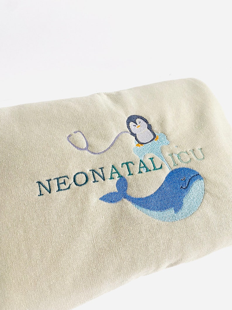 Pode incluir: Um cobertor bege claro bordado com as palavras "NEONATAL ICU". O design inclui uma baleia azul, um pinguim com um jaleco m&eacute;dico e um estetosc&oacute;pio. O bordado &eacute; em tons de azul e verde.
