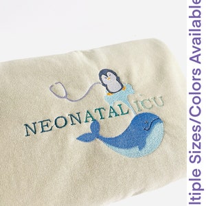 Pode incluir: Um item bordado bege com um pinguim, um estetosc&oacute;pio e uma baleia azul, com o texto "NEONATAL ICU." A imagem tamb&eacute;m inclui o texto "Multiple Sizes/Colors Available."