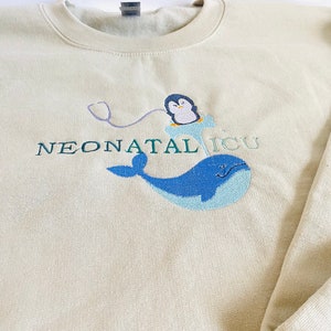 Pode incluir: Camisola creme com design bordado. Apresenta uma baleia azul, um pinguim num icebergue azul e as palavras "NEONATAL ICU" em verde. Um estetosc&oacute;pio roxo liga o pinguim ao texto.