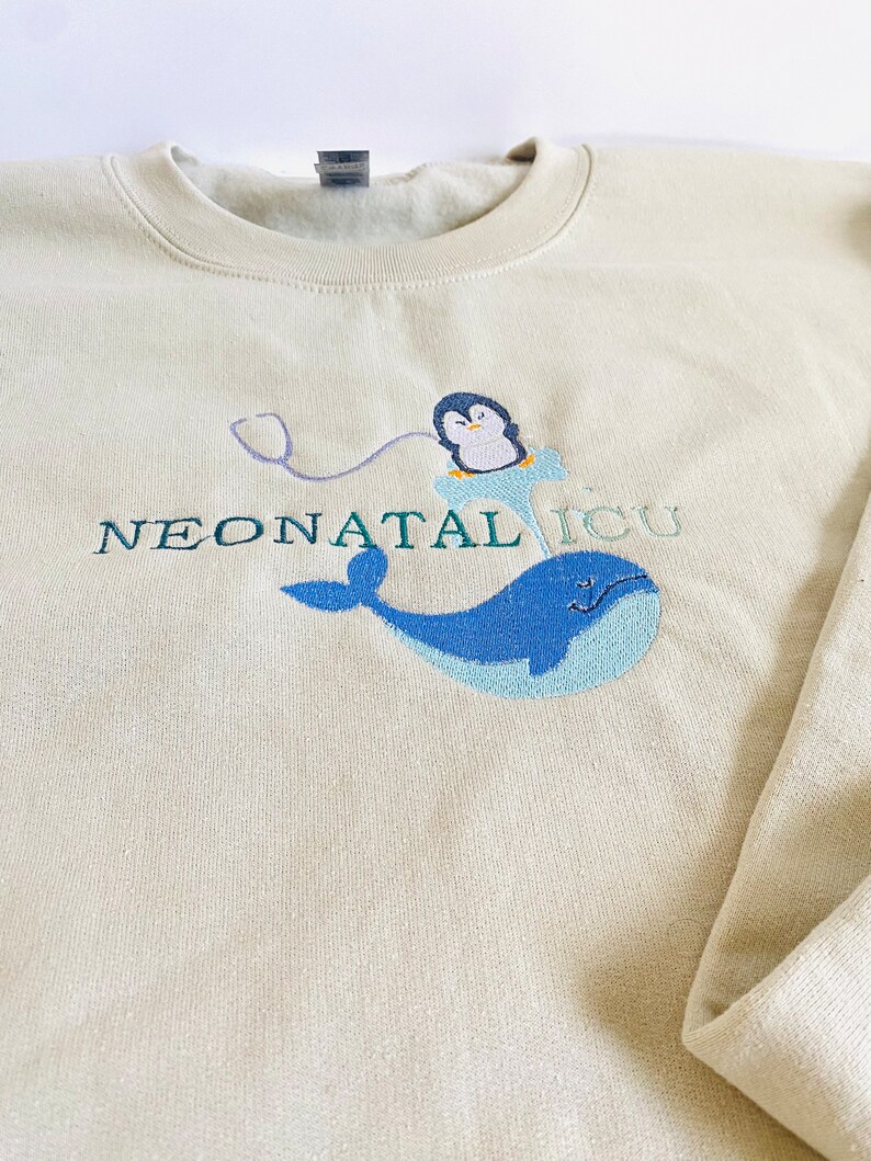 Pode incluir: Camisola creme com design bordado. Apresenta uma baleia azul, um pinguim num icebergue azul e as palavras "NEONATAL ICU" em verde. Um estetosc&oacute;pio roxo liga o pinguim ao texto.