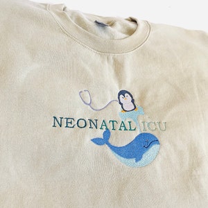 Pode incluir: Um moletom bege com o texto bordado "NEONATAL ICU". O design apresenta um pinguim usando um gorro de enfermeira e um estetosc&oacute;pio, junto com uma baleia azul. O moletom &eacute; feito de um material macio.