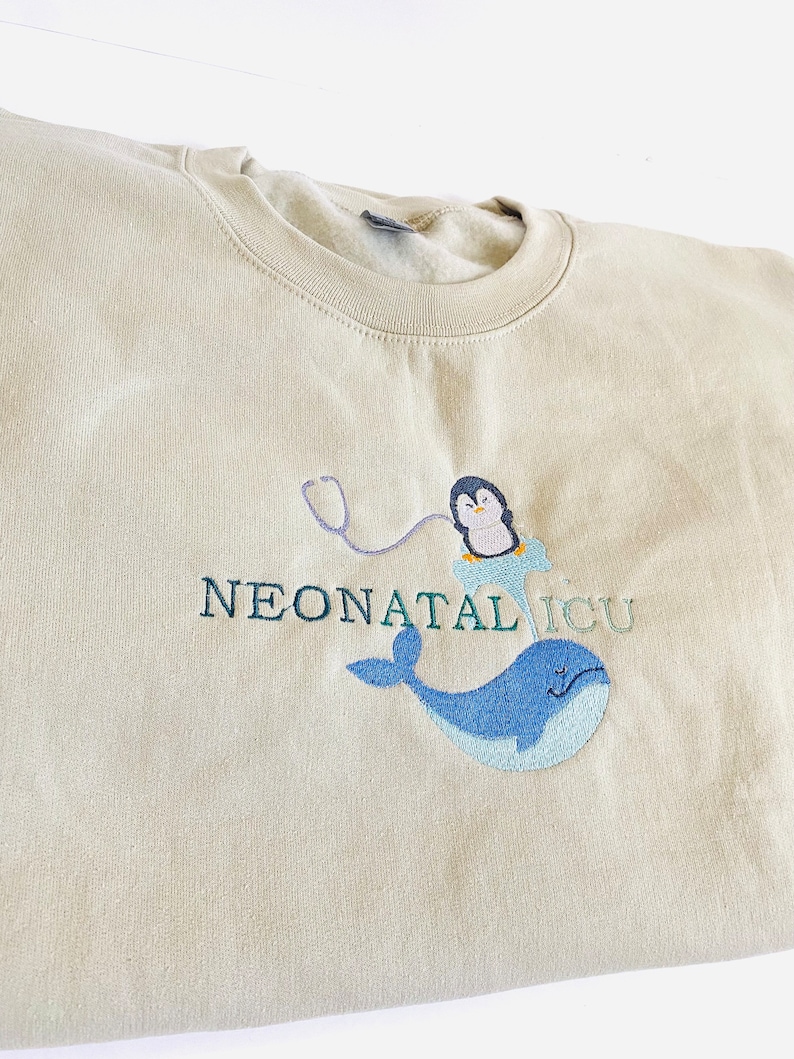 Pode incluir: Um moletom bege com o texto bordado "NEONATAL ICU". O design apresenta um pinguim usando um gorro de enfermeira e um estetosc&oacute;pio, junto com uma baleia azul. O moletom &eacute; feito de um material macio.