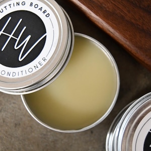 May include: Open tin of cutting board conditioner with a light yellow balm. The tin has a silver lid with the words "CUTTING BOARD CONDITIONER" and a black circle with the letters "HW". The background includes a wooden cutting board.