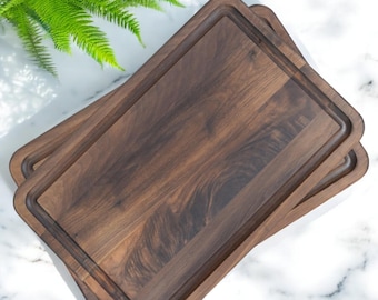 May include: Two rectangular wooden cutting boards with a dark brown finish are stacked on top of each other. Each board has a recessed juice groove around the edge. The boards are on a white marble surface with green foliage in the background.