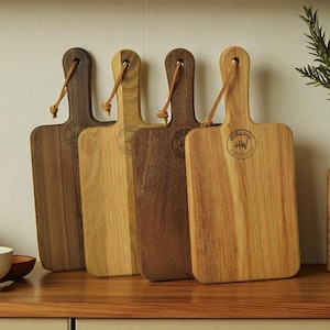 May include: Four wooden cutting boards with handles and leather loops. The boards vary in color from light to dark brown. Each board has a circular logo. A vase with greenery and a bowl with a plate are also visible.