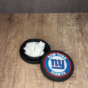 May include: Black, round container with a lid featuring the New York Giants logo in blue, red, and white. The container is open, revealing white packets inside. The container sits on a wooden surface.