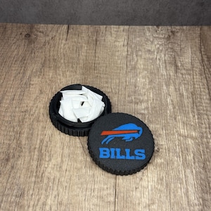 May include: Black, round container with a lid featuring the Buffalo Bills logo in blue and red. The container is open, revealing white packets inside. The lid is off to the side, and the container is on a wooden surface.