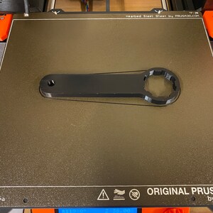Op de afbeelding: Een zwarte 3D-geprinte sleutel met een zeskantige kop, liggend op een getextureerd grijs oppervlak. Het oppervlak is gelabeld "ORIGINAL PRUSA i3 MK3 by Josef Prusa".