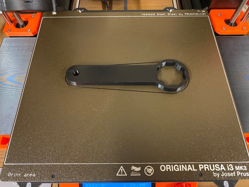 Op de afbeelding: Een zwarte 3D-geprinte sleutel met een zeskantige kop, liggend op een getextureerd grijs oppervlak. Het oppervlak is gelabeld "ORIGINAL PRUSA i3 MK3 by Josef Prusa".