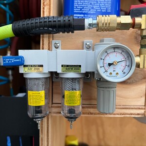 Op de afbeelding: Een close-up van een pneumatisch luchtfilter- en nevelscheidersysteem dat is gemonteerd op een houten oppervlak. Het systeem omvat twee doorzichtige filters met het label "SAF 2000", een manometer met het label "Pneumatic" en een messing elleboogfitting. Het systeem is verbonden met een groene en zwarte luchtslang.