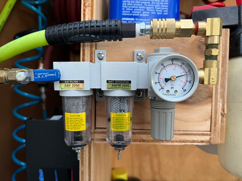 Op de afbeelding: Een close-up van een pneumatisch luchtfilter- en nevelscheidersysteem dat is gemonteerd op een houten oppervlak. Het systeem omvat twee doorzichtige filters met het label "SAF 2000", een manometer met het label "Pneumatic" en een messing elleboogfitting. Het systeem is verbonden met een groene en zwarte luchtslang.