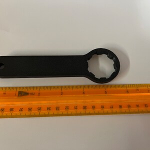Op de afbeelding: Een zwarte kunststof sleutel met een 12-puntige dop. De sleutel is ongeveer 15 cm lang en wordt gemeten met een liniaal.
