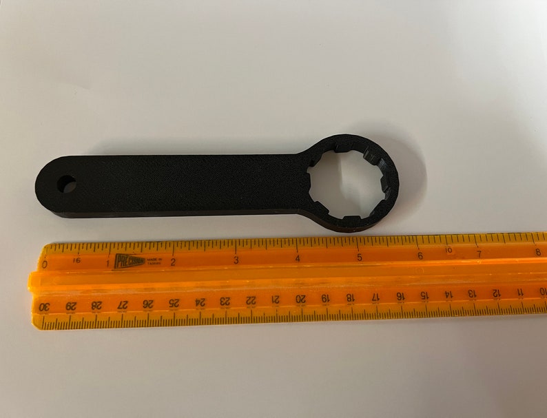 Op de afbeelding: Een zwarte kunststof sleutel met een 12-puntige dop. De sleutel is ongeveer 15 cm lang en wordt gemeten met een liniaal.
