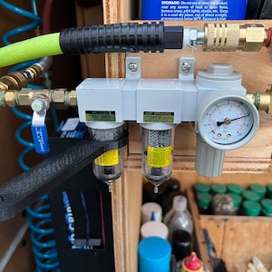 Op de afbeelding: Een close-up van een wit luchtfilter- en waterafscheidersysteem met een manometer. Het systeem is gemonteerd op een houten oppervlak en bevat een zwarte slang en messing fittingen. Het luchtfilter is ge&euml;tiketteerd met "SAP 2000" en de waterafscheider met "SAFM 2000".