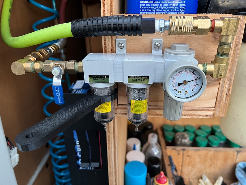 Op de afbeelding: Een close-up van een wit luchtfilter- en waterafscheidersysteem met een manometer. Het systeem is gemonteerd op een houten oppervlak en bevat een zwarte slang en messing fittingen. Het luchtfilter is ge&euml;tiketteerd met "SAP 2000" en de waterafscheider met "SAFM 2000".