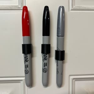 May include: Three permanent markers with black caps and black rings around the barrels. The markers are red, black, and silver. The markers are labeled "Sharpie Fine Point" and have a "AP" symbol.