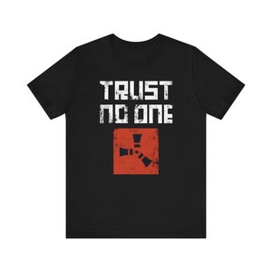 May include: Black t-shirt with the text "TRUST NO ONE" in white, distressed font. Below the text is a red square with a stylized, rust-colored graphic. The shirt is a crew neck style.